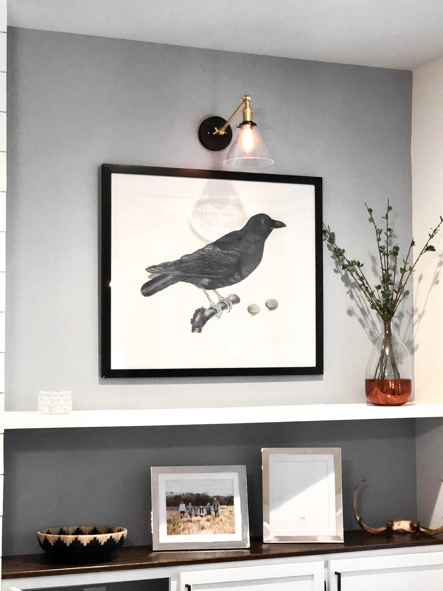 Mid-century modern wall sconce with polished brass finish and adjustable clear glass shade above framed black bird artwork. - SCONCE MODEL NO. 7962