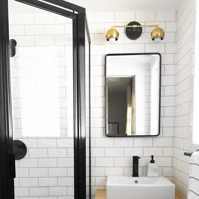 Mid-century modern 2-bulb vanity light with black and raw brass finish above rectangular mirror in white tiled bathroom. - VANITY LIGHT MODEL NO. 5469
